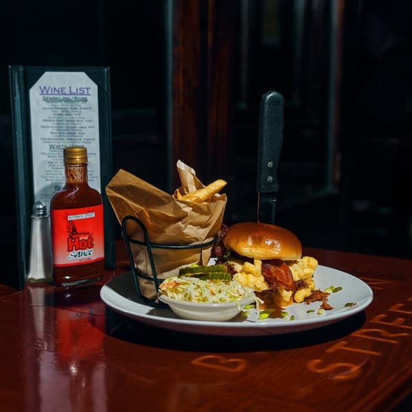 Bourbon Street's mac and cheese burger with fries, coleslaw, and hot sauce on a dark wooden table.